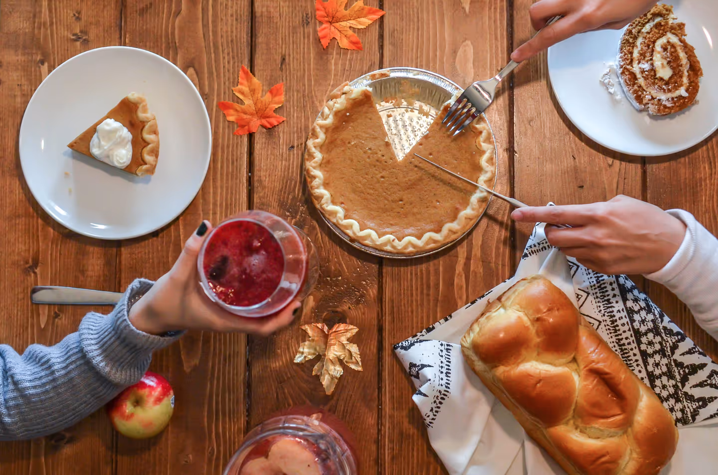 A wooden table seen from above with pumpkin pie being sliced, a braided loaf, a pumpkin roll and a glass of cranberry punch, hands reaching in from both sides